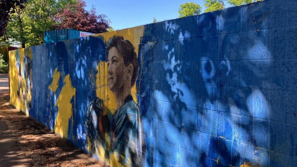 A stretch of brick wall spray-painted blue and yellow and featuring the figure of a boy holding a football.