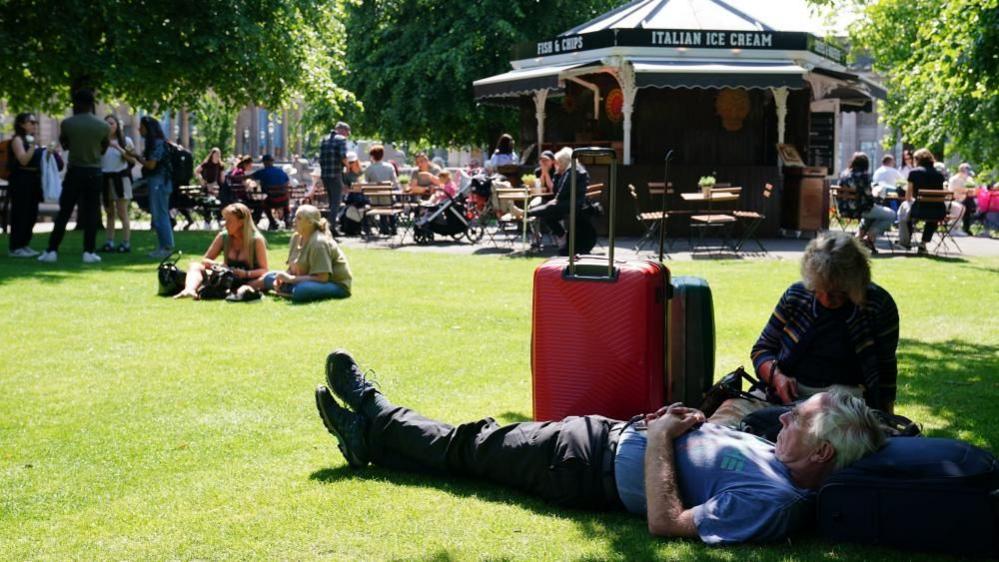 A man reclines in the shade on grass in front of a pavilion selling ice cream in Edinburgh's Princes Street Gardens.
