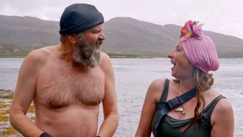 Greg and Jules look at each other and seem to be laughing in their swimming gear. Jules is wearing a pink headwrap with flowers at the front and Greg is wearing a grey beanie hat. It is a grey day and they are standing in front of water with hills in the background.