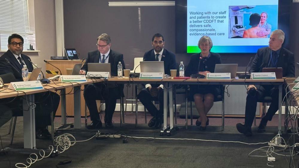 Four men in suits and a woman in a skirt and blazer sit around long desks in a room. On a screen behind them there is a picture of a nurse touching a patient's back and some black text.