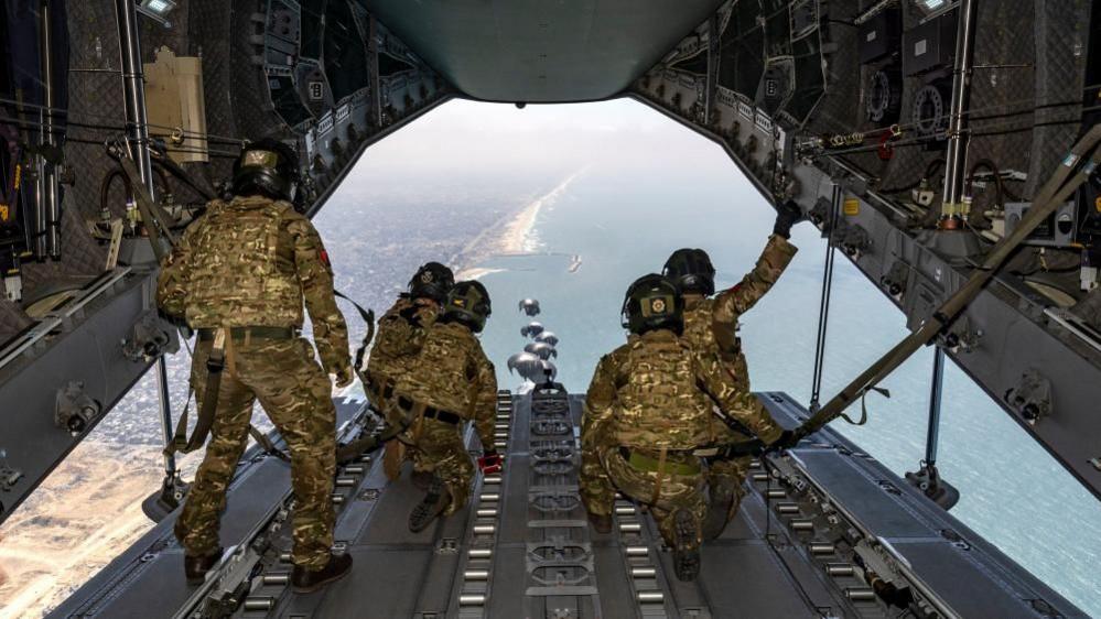 Airmen in khaki uniform watch from the open rear of an airplane as supplies are dropped by parachute 