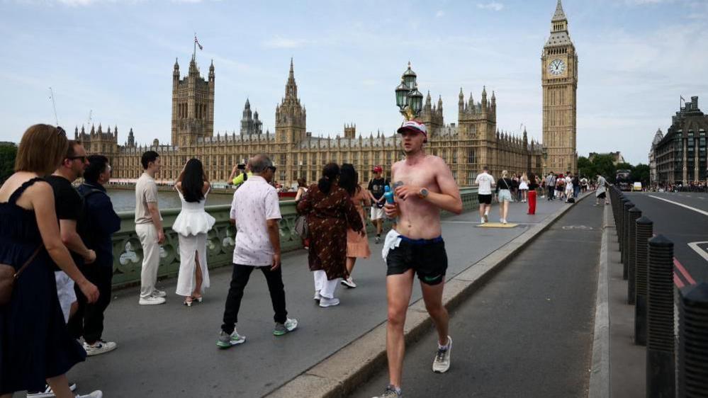 A man jogs across Westminster Bridge towards the camera with other people walking across in the opposite direction. The Houses of Parliament can be seen in the background.