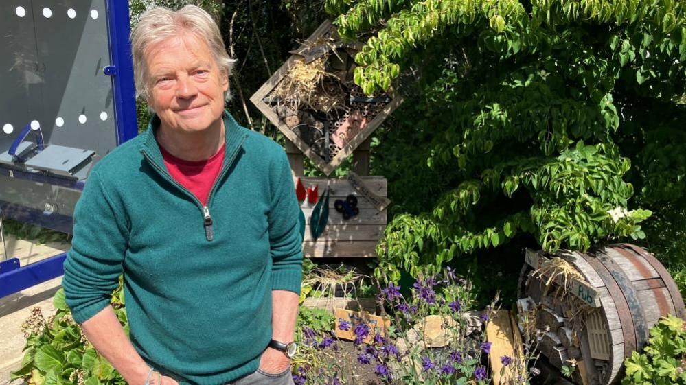 A man in a teal-green jumper with red T-shirt underneath stands in front of two wooden bug hotels, smiling at the camera. A railway station shelter can be seen to his left.