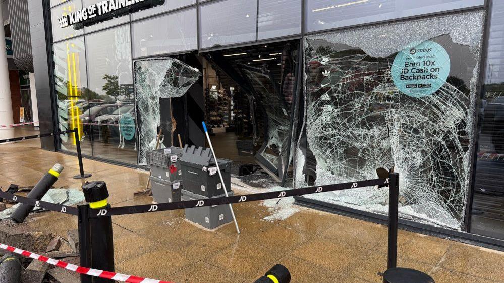 Another angle of the damage. The door has been warped. Grey boxes sit in front of the hole in the glass front. Brooms and poles can also be seen.