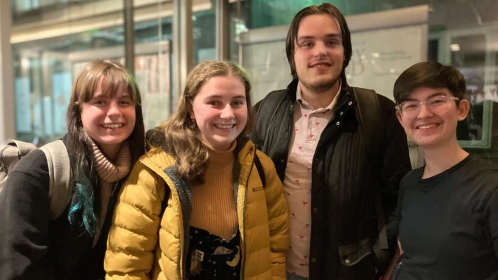 Four people stand wearing coats on their way out of a cinema. One is wearing a pink shirt while another has green hair.