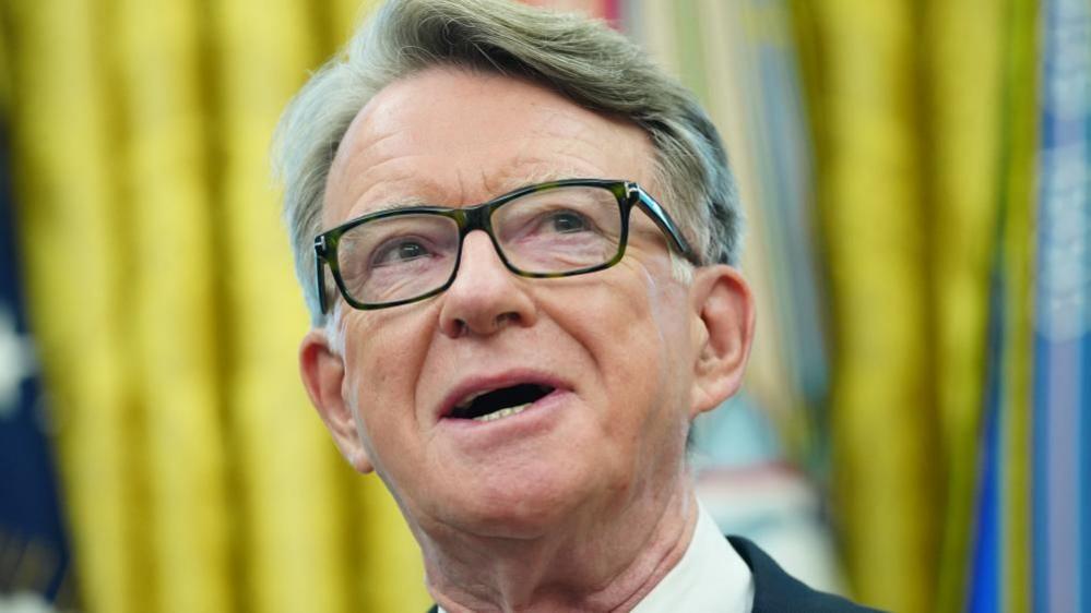 A close-up image of a man, Peter Mandelson, wearing glasses with grey hair in a quip, his mouth open while talking in front of gold curtains.
