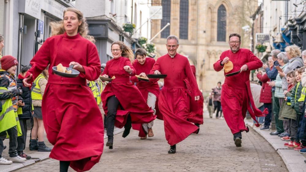 Two women and three men in red cassocks run down a street holding frying pans with crepes in them. 