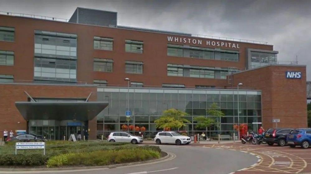 The main entrance to Whiston Hospital can be seen in this image from Google. There are a number of cars parked outside and there is a large glass panelled area at the front.