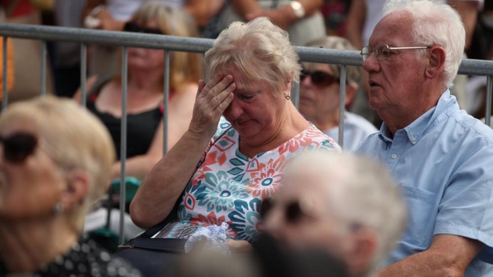 An older woman with short grey hairs looks visibly upset, with her right hand raised to her forehead and her head bowed. A man with glasses and short grey hair is sitting next to her and blurred out people can also be seen around them.