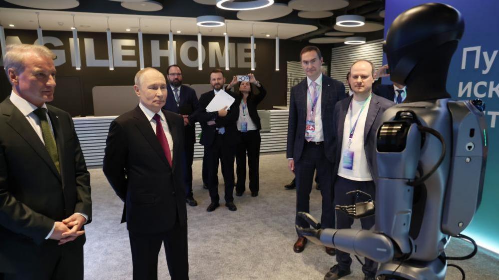 Putin, wearing a black suit with a red tie, looks at a robot infront of him. He is expressionless.