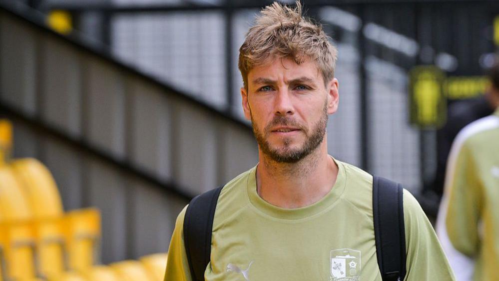 Sam Foley arriving for a Barrow match wearing a pale green t-shirt with the Barrow badge on the left against a blurred backdrop of yellow seats