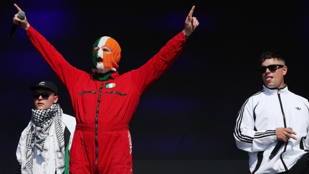 Left to right: Kneecap's Mo Chara, DJ Provai, and Moglai Bap stand on stage at Glastonbury