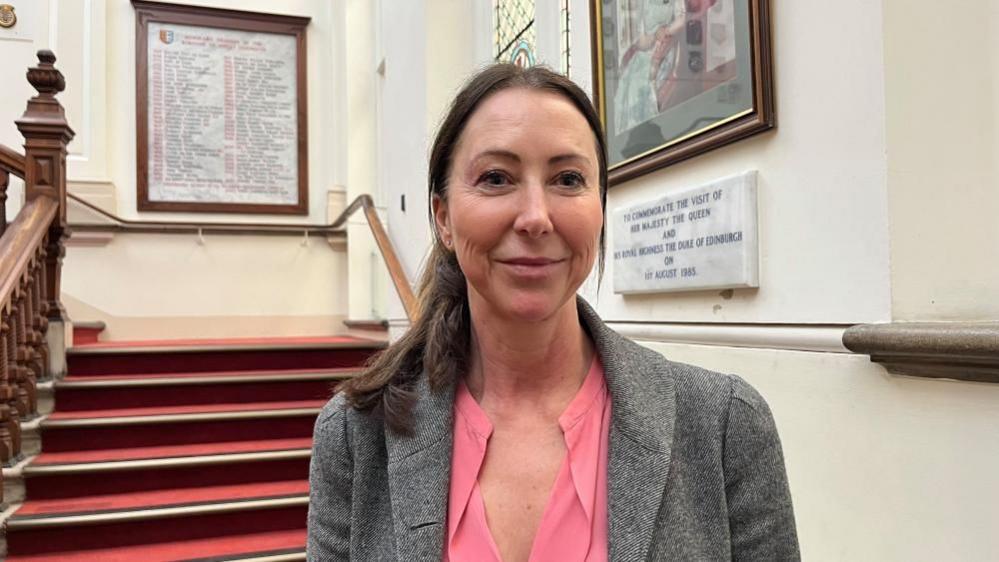 Sheila Oxtoby is standing on the staircase at Great Yarmouth town hall, with marble tablets and portraits hung on the walls. She has long dark hair, tied back, and is wearing a pink blouse with a grey jacket over.