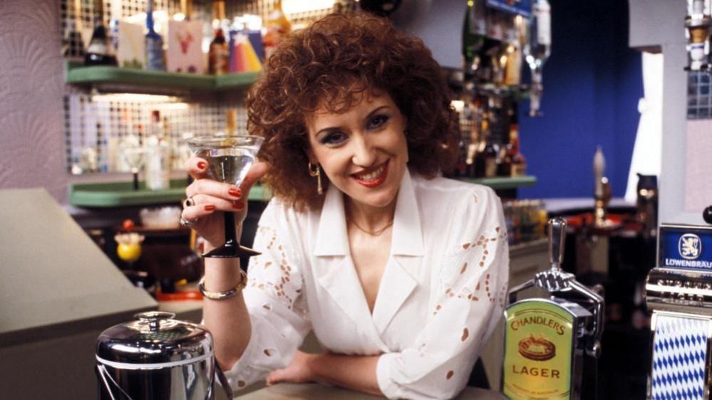 Anita Dobson dressed as Angie Watts while she was on the show in the 1980s. She is standing behind a bar smiling and holding a martini glass.