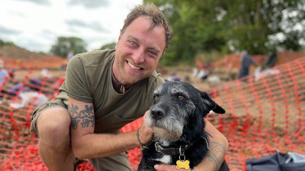 Oliver wearing a green t-shirt kneeling down on the excavation site next to his black dog Nero