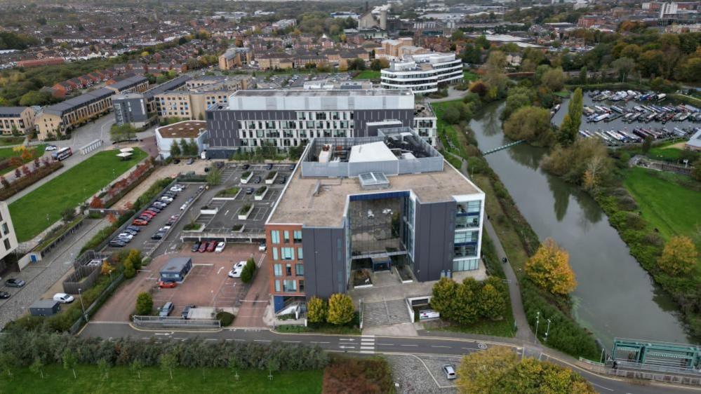 Aerial shot of the UON Waterside Campus, showing river to the right, and modern grey and white university buildings to the left.  There are houses in the background.