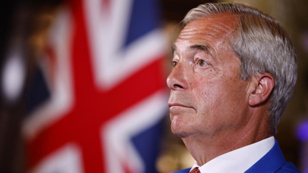 Nigel Farage is wearing a blue suit, white shirt and a tie. He has short grey hair. A Union flag can be seen in the background.
