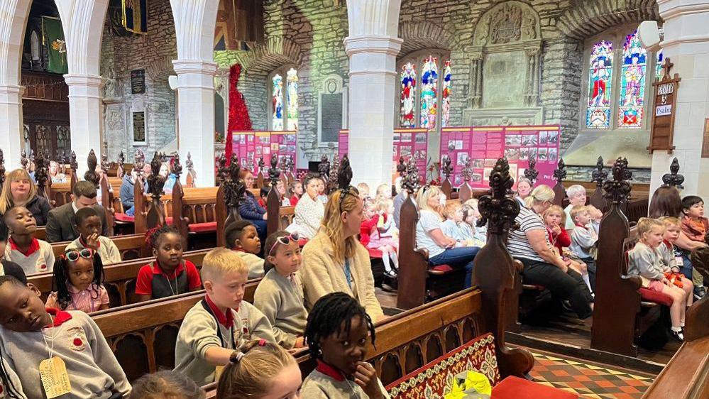 A large crowd of schoolchildren in grey uniforms, on wooden benches inside a cathedral, there are white beams and stone walls