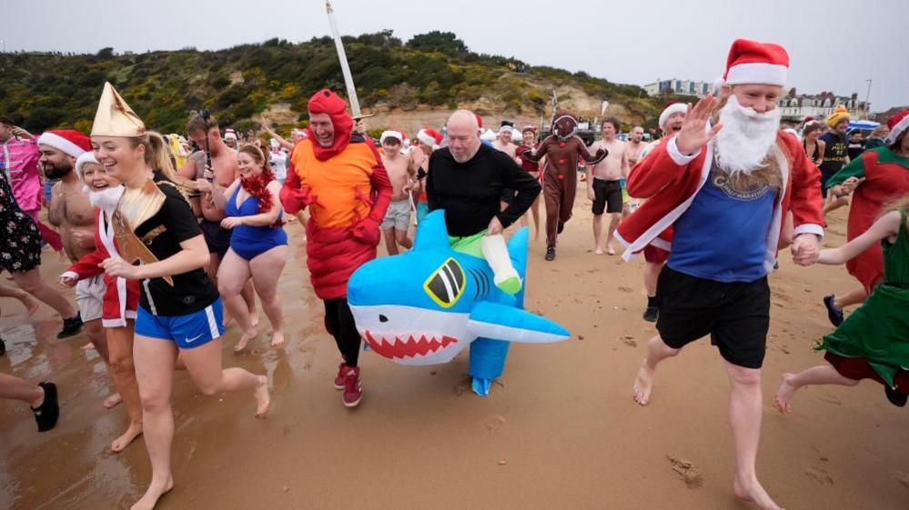 A man wearing a shark inflatable costumer runs down the beach with others in fancy dress