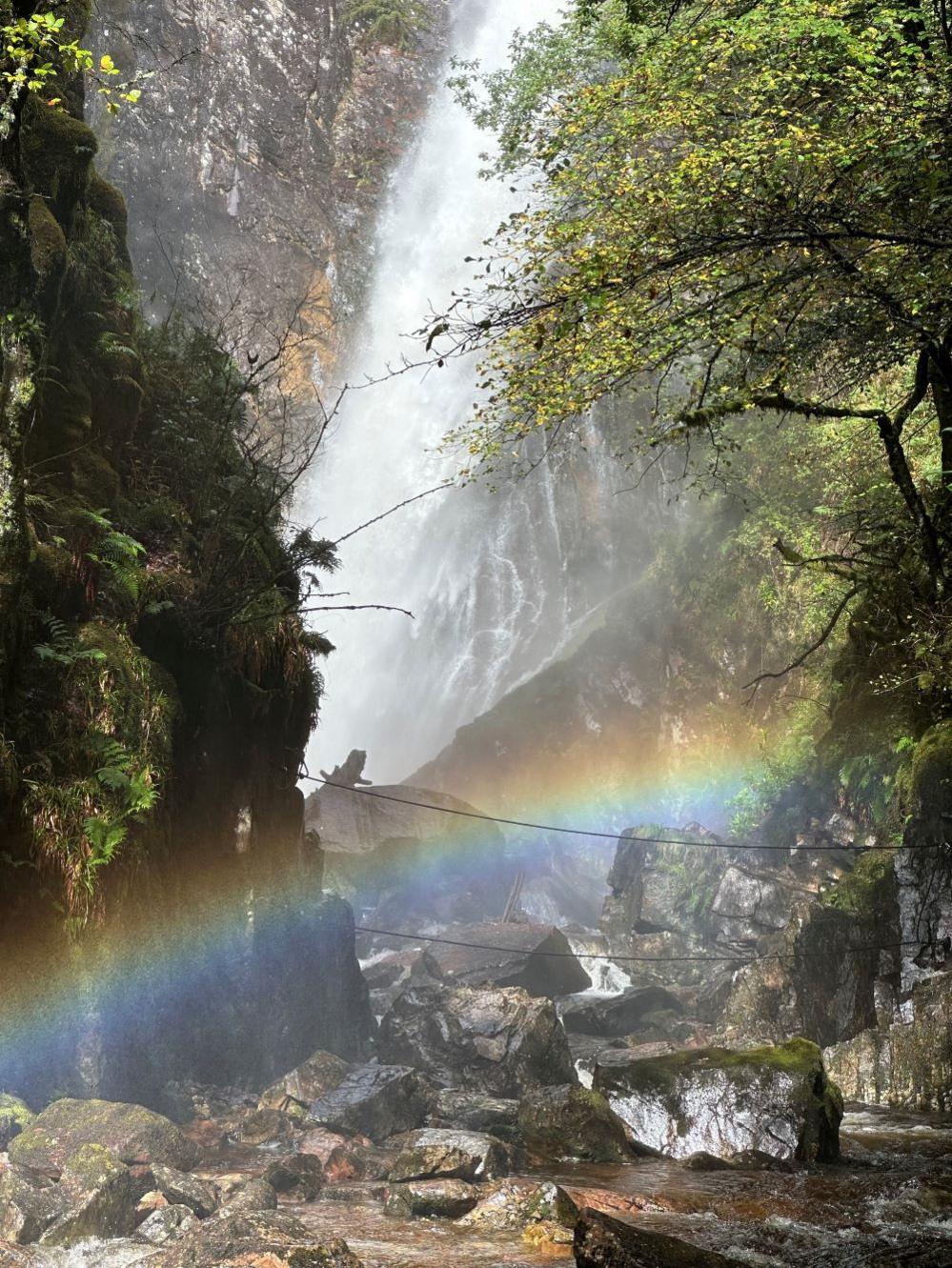 A vibrant rainbow arches across a rocky stream bed, cascading down from a powerful waterfall.