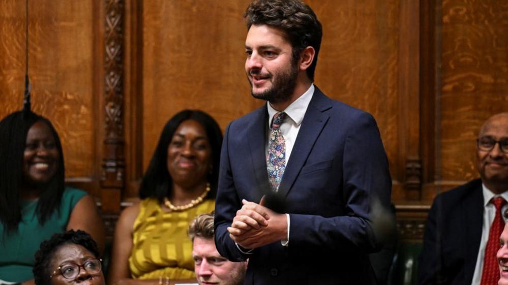 MP Jack Abbott standing in House of Commons with other MPs looking on. He is wearing a dark suit, white shirt and and multicoloured ties with stars on it