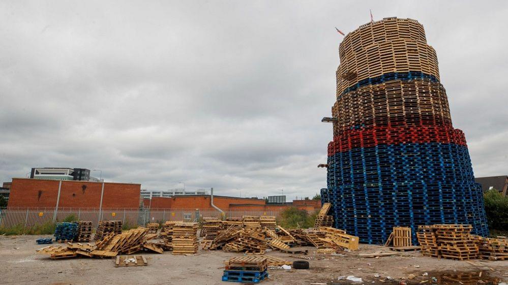 The south Belfast bonfire with red and blue pallets and brown ones. There are pallets scattered on the ground beside the bonfire that stands taller than the surrounding buidlings. 