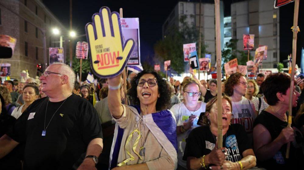 A woman in the crowd holds up a poster of a yellow hand that says 'bring them home now'