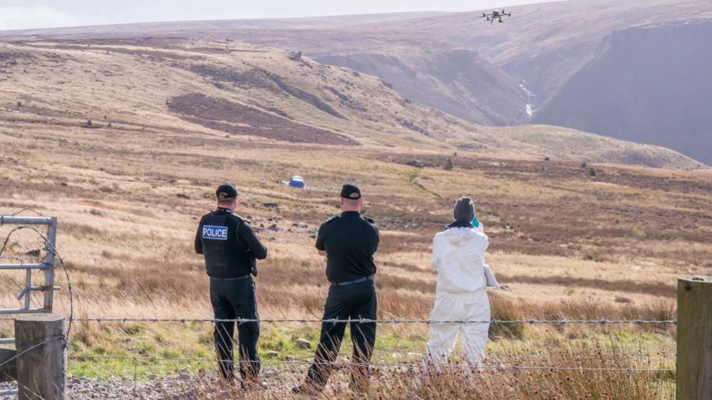 Three police officer - two in uniform and one in a forensic suit - look out over a moor. A blue tent can be seen set up in the distance.