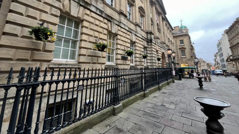 An exterior view of the market building. It is a historic bath-stone structure, with Georgian windows, and metal railings across the front. In the foreground, a brass "nail" can be seen, which was formerly used as a trading platform