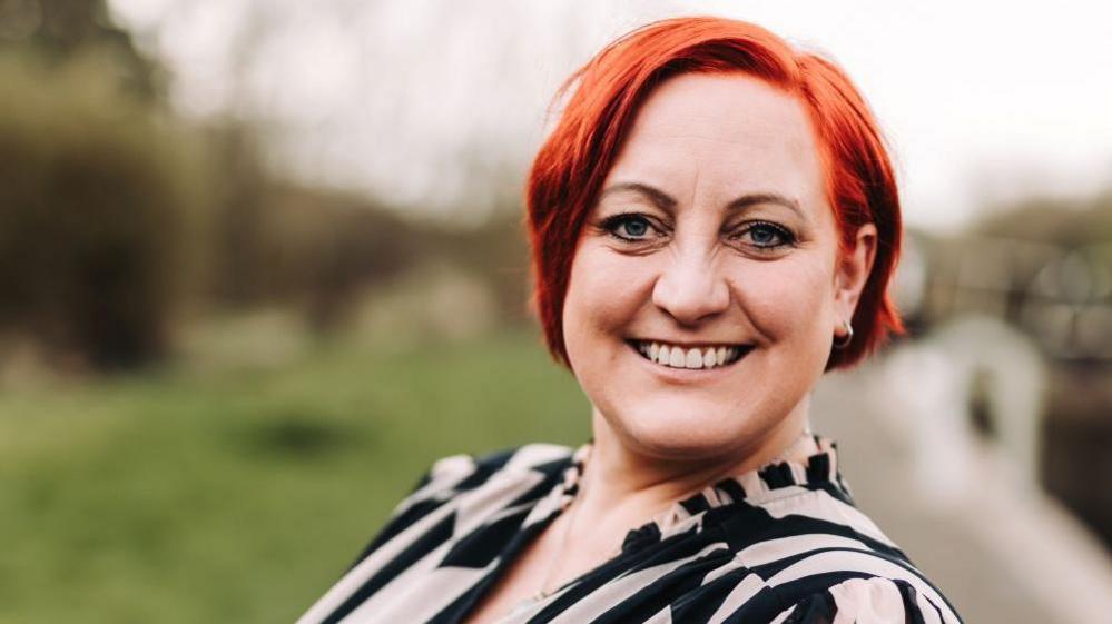 A smiling Esther Underwood looks directly at the camera as she photographed outside. She has red hair and is wearing a patterned top. 