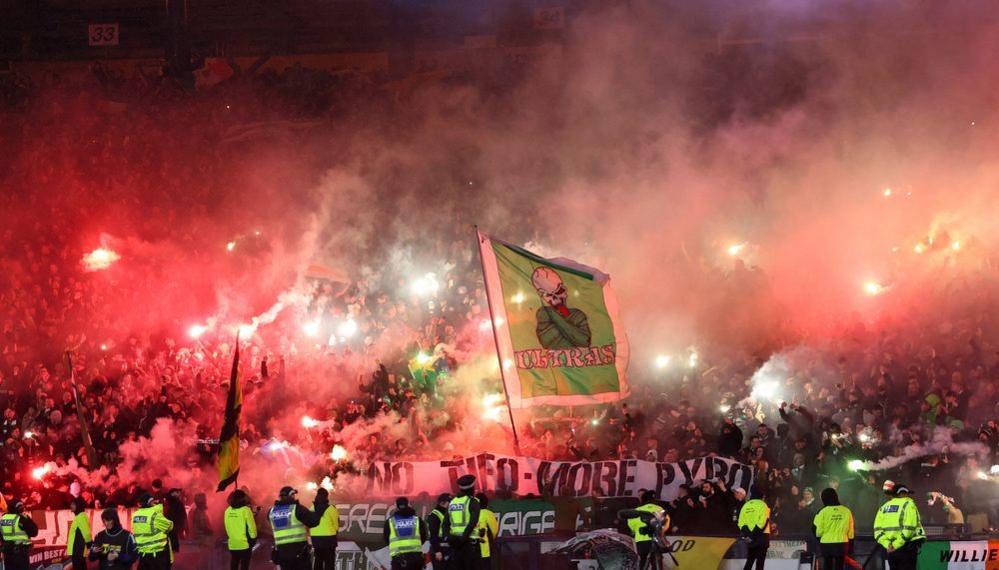 Smoke and red flares visible in a stand full of football fans. Multiple stewards and police officers wearing high vis jackets are visible. A large flag emerges from the crowd. It is green and with a skeleton on it.
