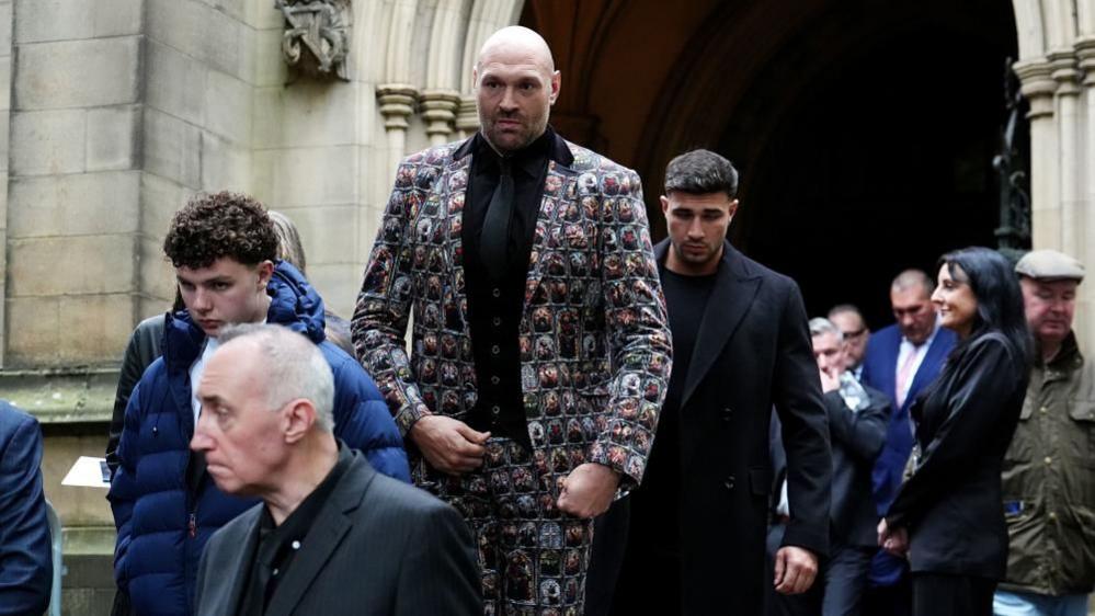 Tyson Fury wearing a colourful suit walks out of a church. Tommy Fury, wearing a black suit, walks behind him.