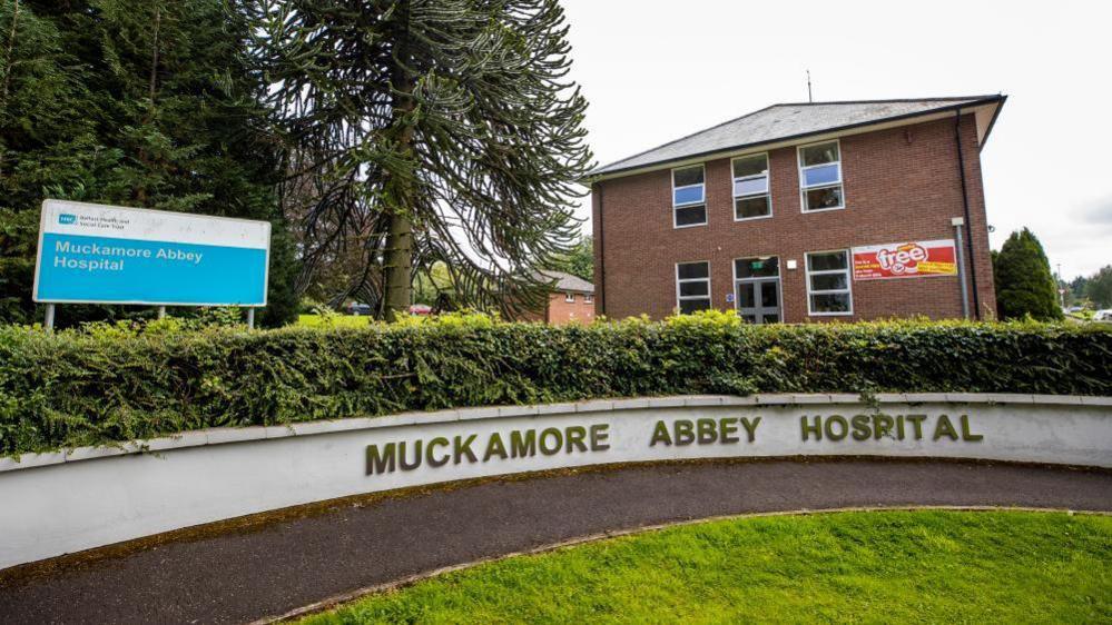 A sign that says Muckamore Abbey Hospital in gold writing on a white wall. 