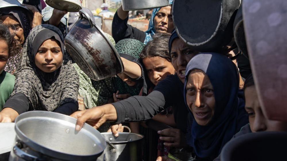 Palestinians queue to receive a meal from a kitchen that provides free food for displaced people in Khan Yunis, they are holding out empty pans and crowded closely together