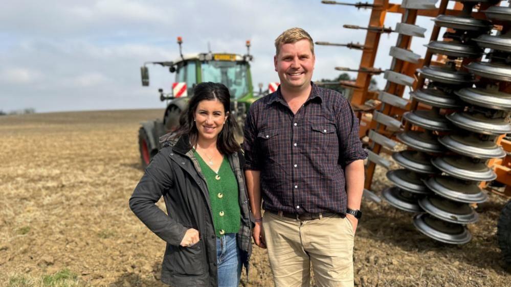 Kate Scott wearing a dark green coat, green top and blue jeans is standing next to Lewis Hunter, who is wearing a dark check shirt and beige trousers. Behind them is a large combine harvester and a ploughed field.