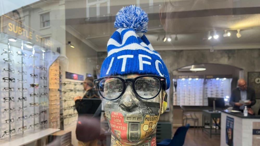 An optician's mannequin wearing blue and white bobble hat 