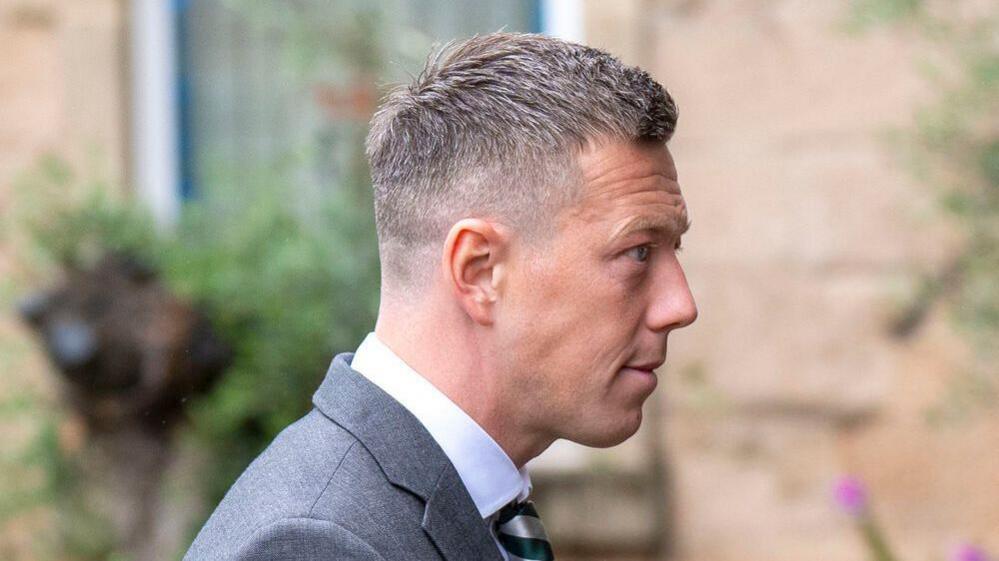 A side profile shot of a man with short brown hair wearing a grey suit