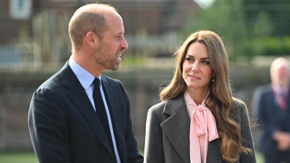 A picture of Prince William in a navy suit jacket with navy blue tie and blue shirt talking as Princess Kate, wearing a grey overcoat and pink-bow blouse listens in. 