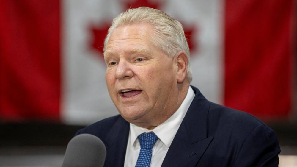 Premier Doug Ford speaks into a microphone during a campaign stop at Walker Construction in Niagara Falls. There is a Canadian flag behind him 