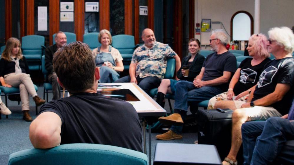 Eight people sitting in a semi-circle of blue chairs in front of brown wooden and glass doors in front of  composer Liam Taylor-West. He is sitting with his back to the camera with dark, short hair and a blue T-shirt.