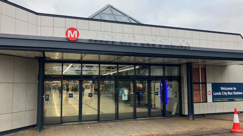 The entrance to a building which has a modern design with light-coloured panels and a triangular glass roof section. Above the glass doors, the sign reads ‘Leeds City Bus Station’.