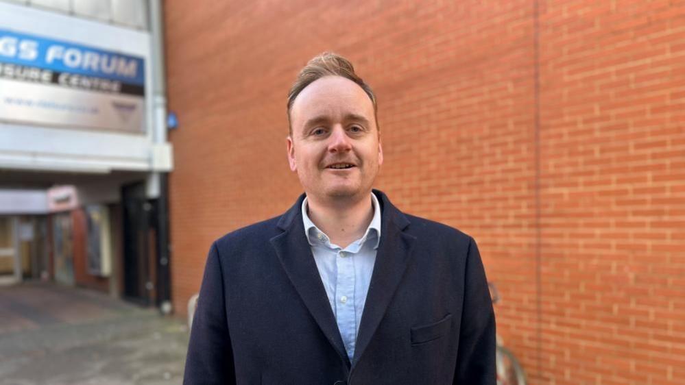 Adam Brown wearing a blue shirt and blue jacket. Is standing in front of an orange wall with a sign for a leisure centre over his shoulder.