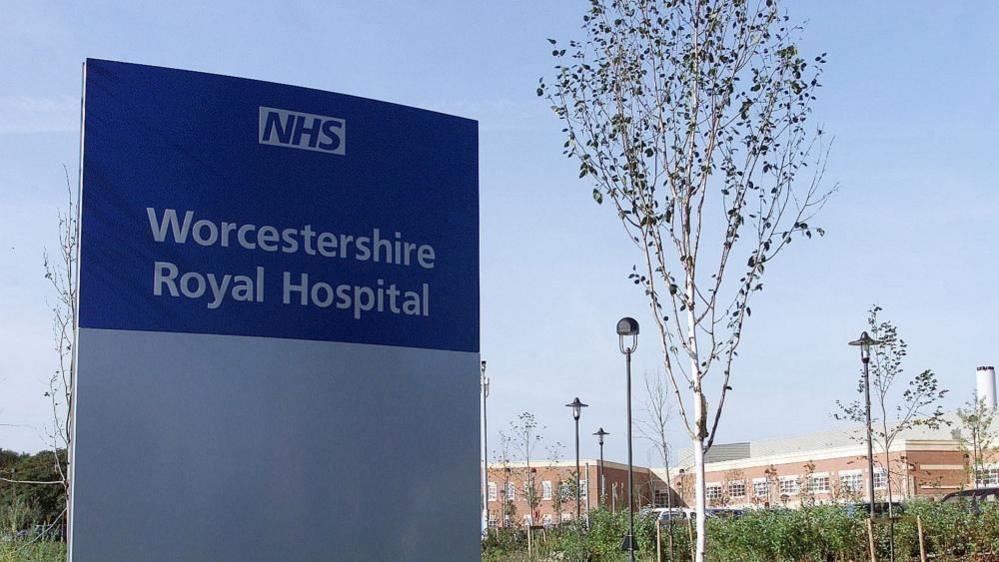 A picture of an NHS hospital sign that reads "Worcestershire Royal Hospital". The red brick hospital building and thin trees can be seen in the background.