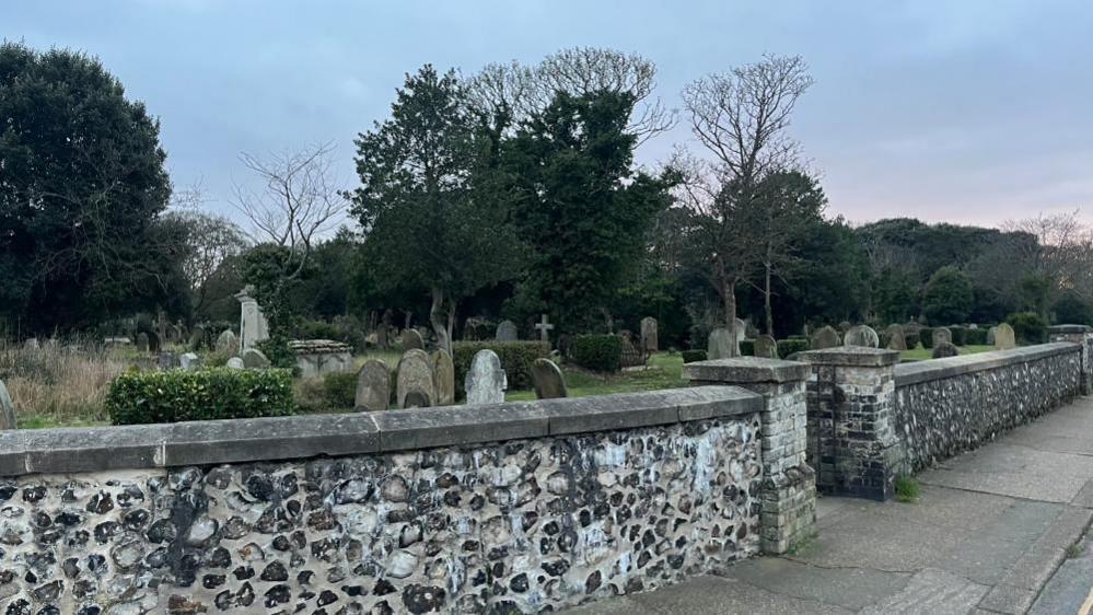 A flint wall about 1m (39 inches) high, built of knapped flint with stone coping. Beyond it are many grave stones, and trees. The sky behind is blue with a tinge of the pink sunset towards the right of the image.