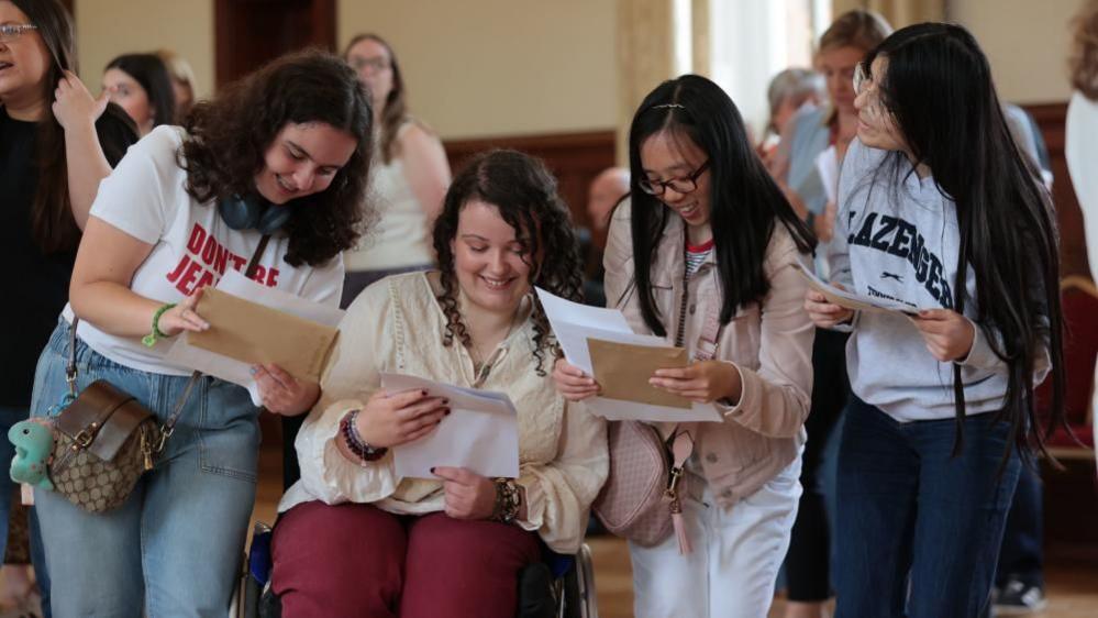 Four female pupils read the papers in their hands and smile looking down. 