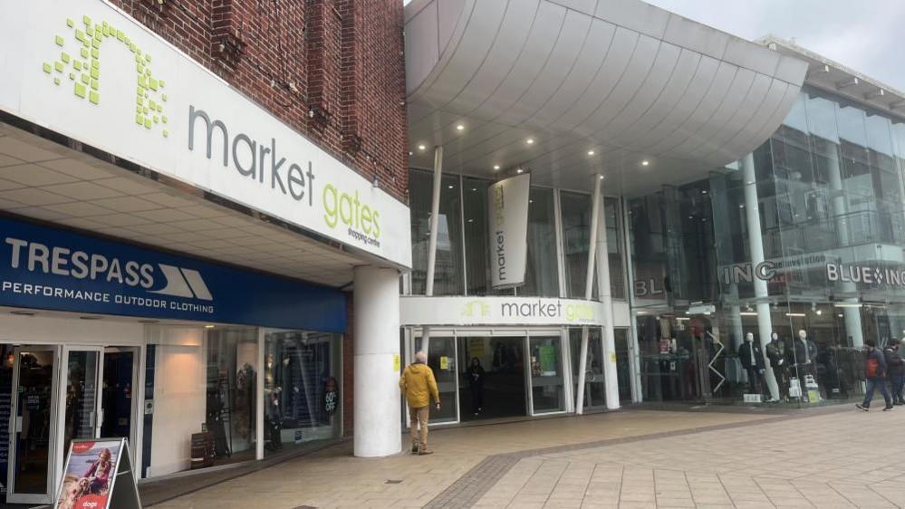 Market Gates shopping centre, with branding in lime green and grey on a white background. The shopping centre features the Trespass shop in the nearground, and Blue Inc, housed within a glazed unit on the right of the main entrance that has a white canopy above.