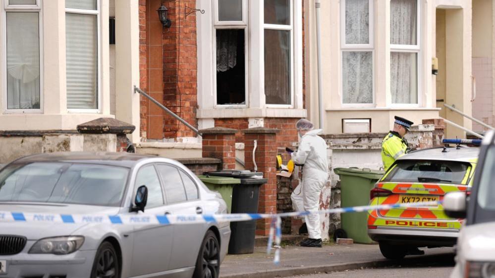 A terraced house in red brick with white window frames. A person in white overalls is at the entrance to the path leading to the door of the house. A police officer in yellow high-vis is also visible, standing next to a police car. Police tape is stretched across the road.