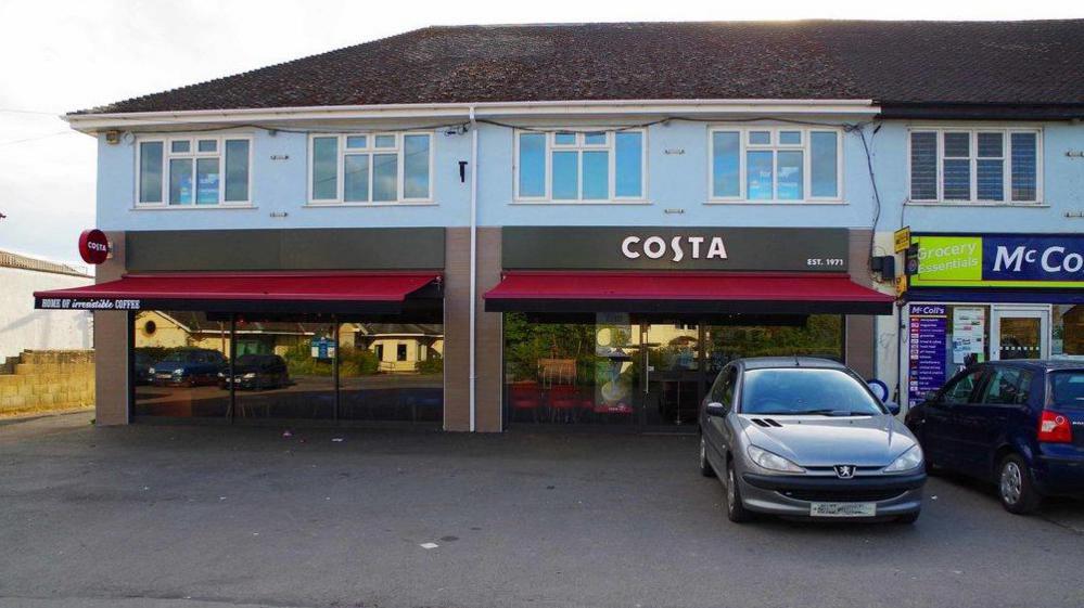 The front of the coffee shop, which has floor to ceiling glass windows. Cars are parked on a forecourt.