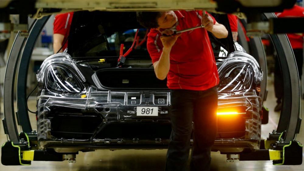 A man in a red tshirt uses a tool on the underside of a car, while a car in the process of being built sits behind him in a factory in Germany.