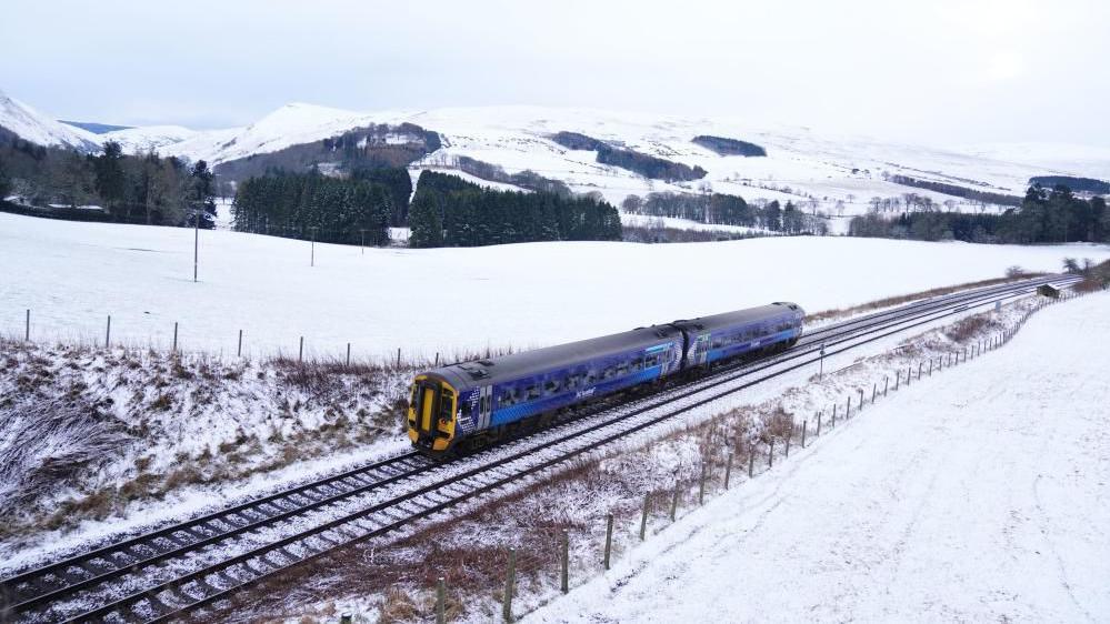 A blue train travelling along a snowy track, surrounded by snow-covered fields in Scotland.